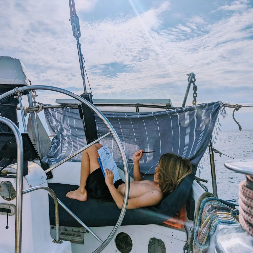 A child is laying and doing his homework on a boat while a black Wetcat Turkish towel is tied in a way to block the sun adn create him a sun share