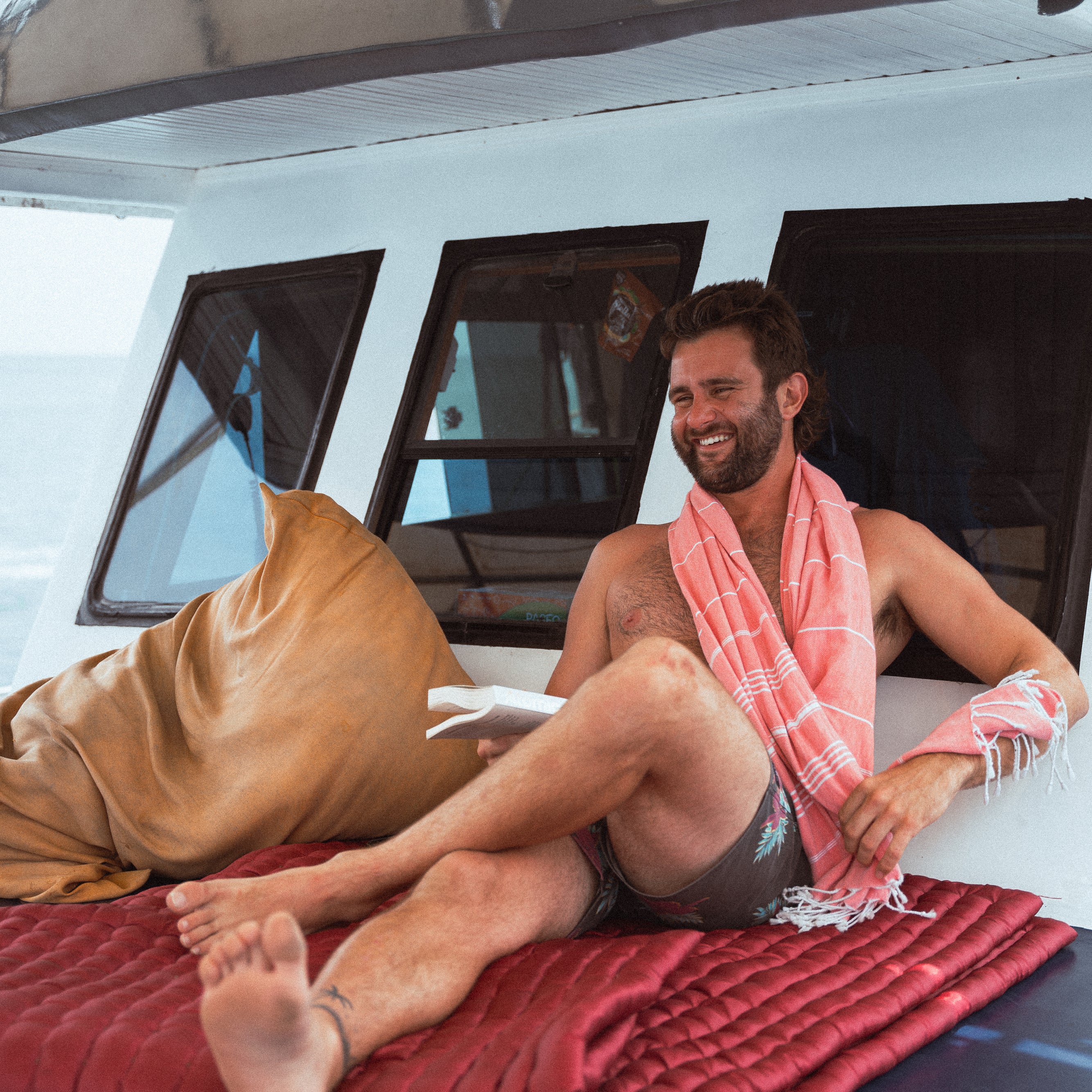 A guy has a dark coral Wetcat Turkish towel wrapped around his neck while sitting and reading a book on a boat