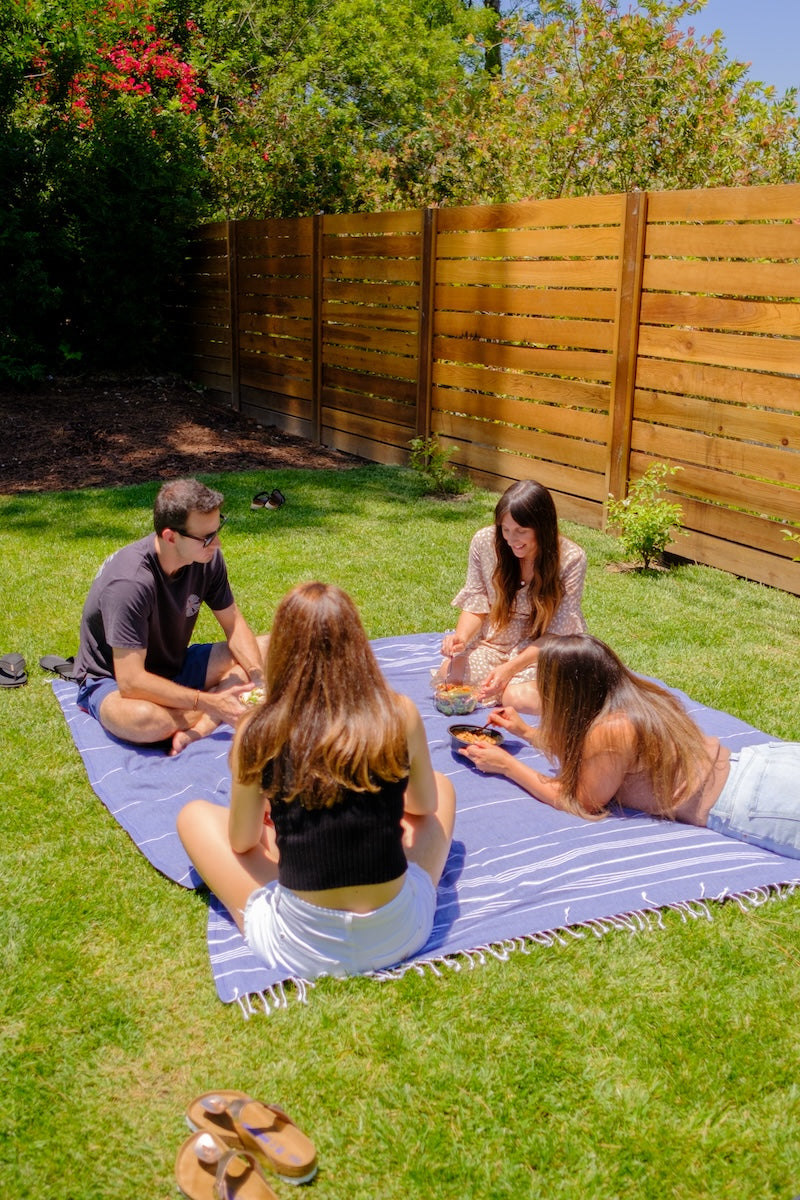 A big navy Wetcat Turkish blanket and 4 people are sitting on it on grass in a backyard